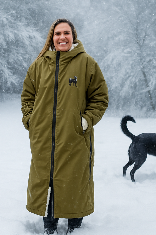 “Woman wearing a long waterproof fleece-lined forest green dog walking coat standing in heavy snow with a winter forest background.”