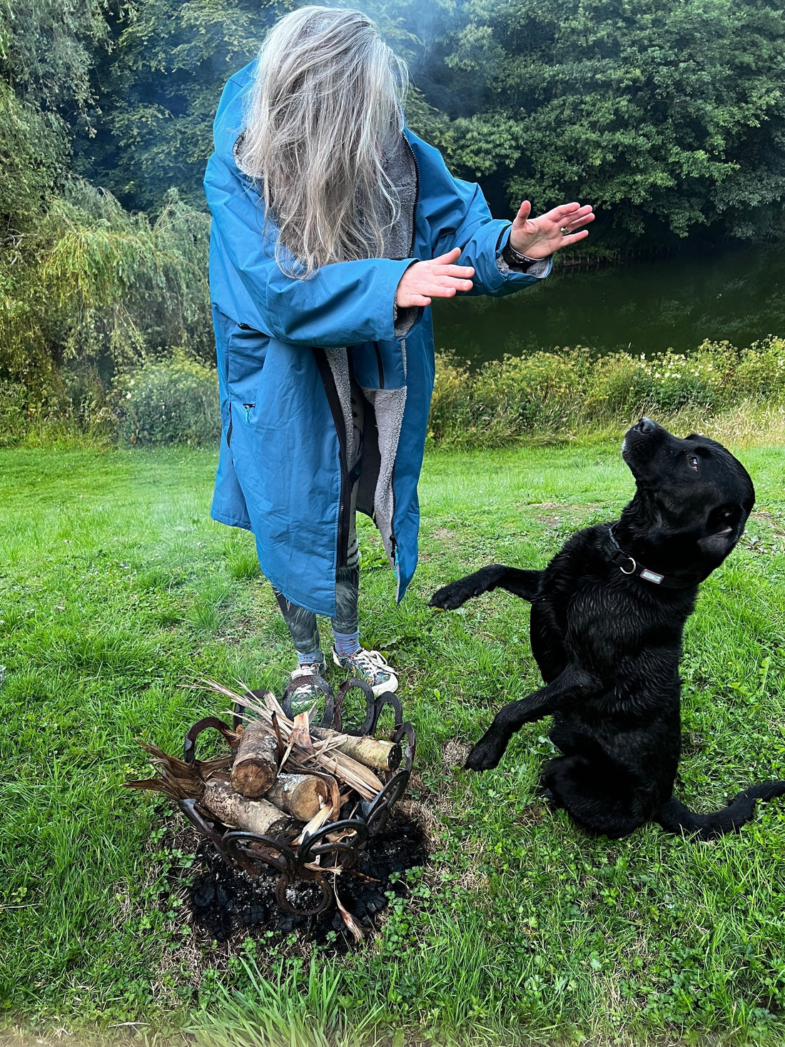 Woman in a long fleece-lined waterproof coat standing with her dog by a bonfire on a cool evening